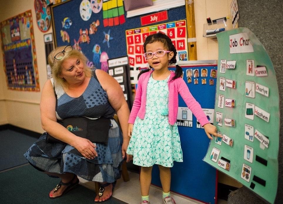 Pre-school girl in glasses standing at an activity board while teacher looks on.